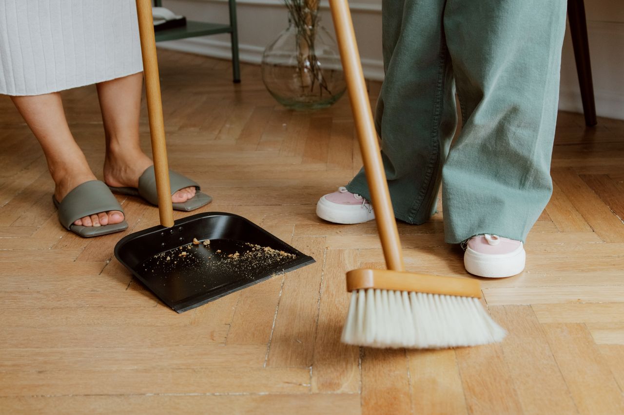 Gros plan sur des personnes balayant un sol en bois à l’intérieur avec un balai et une pelle à poussière.